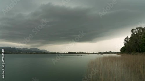 Grey storm clouds pass across white sky over lake edge with wall of rain in wetzikon, Switzerland, wide angle photo