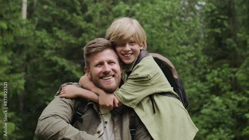 Medium slowmo portrait of cheerful Caucasian boy embracing his dad and both smiling at camera while having family camping trip in woods at daytime Medium slowmo portrait of cheerful Caucasian boy embracing his dad and both smiling at camera while having family camping trip in woods at daytime
