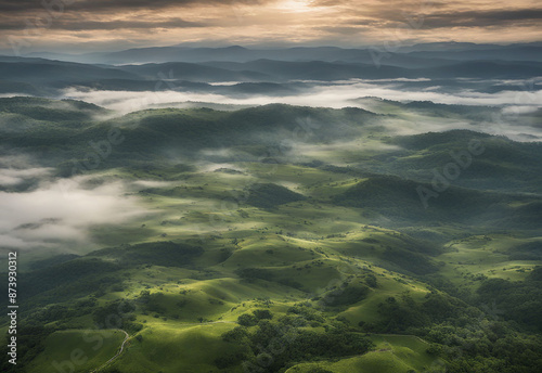 This stunning aerial view captures the ethereal beauty of a mist-laden valley. Rolling hills cloaked in lush greenery stretch out beneath a sky painted with dramatic clouds. The soft mist weaves throu photo