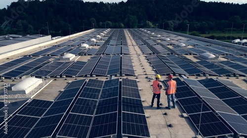 Drone rising above two contractors standing on rooftop solar panel array. Workers in safety helmets walking inspecting sustainable energy PV modules aerial back view