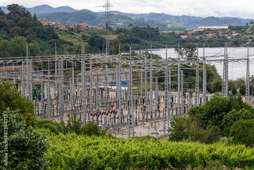 An image of a large electrical substation filled with numerous transformers and complex electrical lines, set against a backdrop of lush, green hills and power lines.