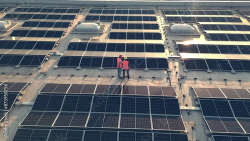 Aerial shot of engineers in helmets walking across photovoltaic array on rooftop. Electricians inspecting solar panels electrical safety at sunset rotating drone shot