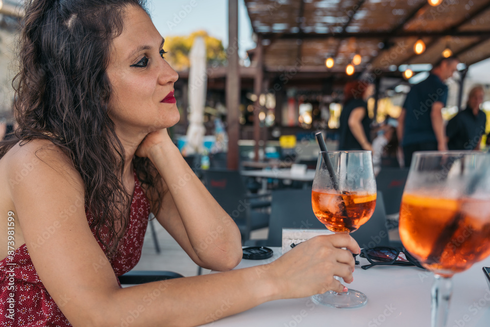Young woman holding spritz cocktail glass sitting at restaurant table. Young woman is sitting alone at a table, holding a spritz cocktail glass and looking away
