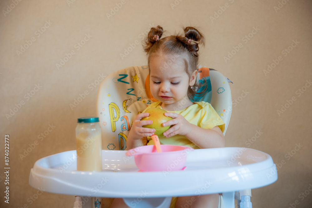 A little girl is sitting on a high chair and eating a green apple at home life style A little girl is sitting on a high chair and eating a green apple at home life style