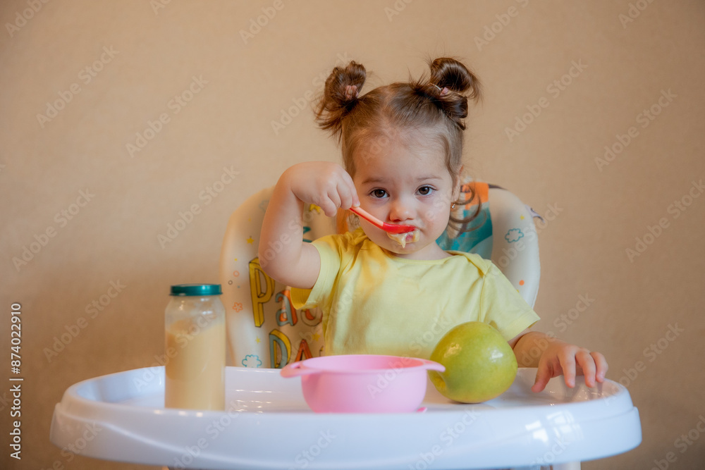 A little girl is sitting on a high chair and eating mashed fruit at home A little girl is sitting on a high chair and eating mashed fruit at home