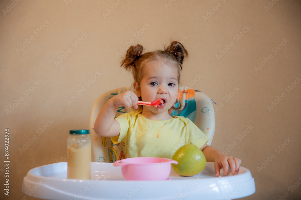 A little girl is sitting on a high chair and eating mashed fruit at home A little girl is sitting on a high chair and eating mashed fruit at home
