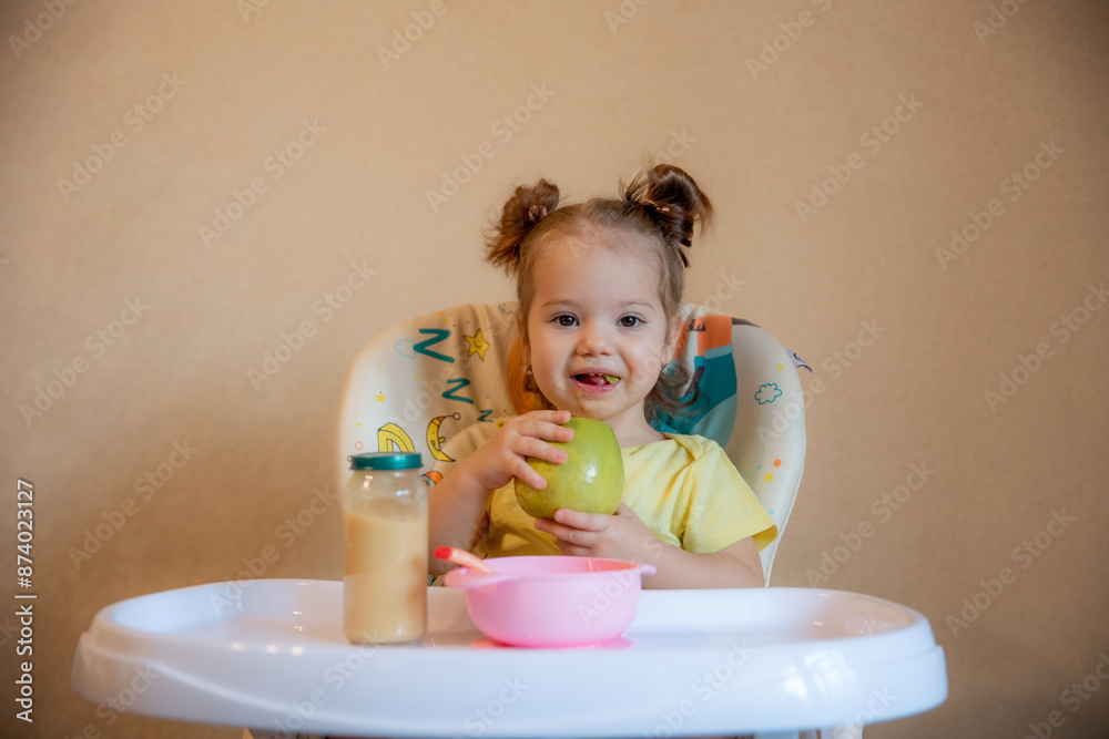 A little girl is sitting on a high chair and eating a green apple A little girl is sitting on a high chair and eating a green apple