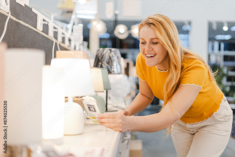 Smiling young woman holding lamp while choosing in decor department in the shopping center Smiling young woman holding lamp while choosing in decor department in the shopping center