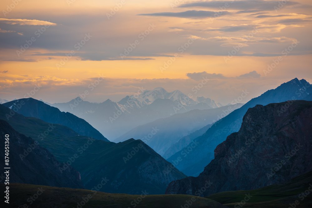 Fototapeta premium Deep gorge between layered silhouettes of sharp rocky ridge spurs under huge snowy mountain peak in golden cloudy sky. Sheer crags of mount valley under giant ice top under gold sunset color clouds.