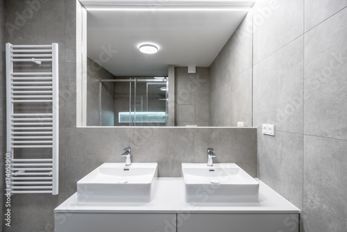 A modern bathroom with gray cement tiles, a double sink with chrome faucets, a large mirror, a glass-enclosed shower, a wall-mounted towel heater, and LED strip lighting. photo