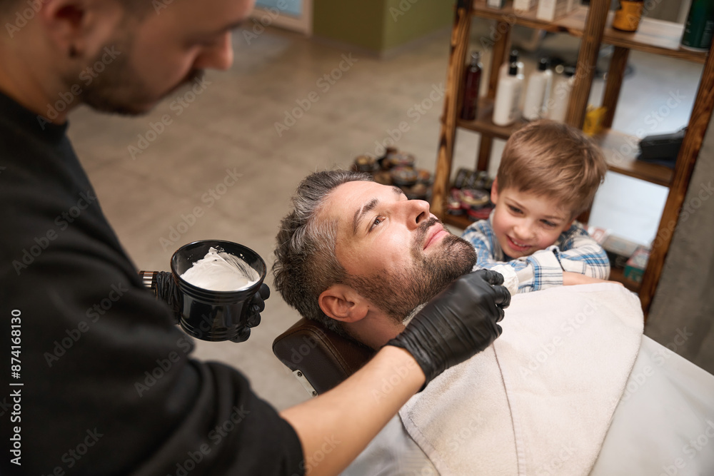 Curious boy watching barber applying shaving cream on client's neck Curious boy watching barber applying shaving cream on client's neck