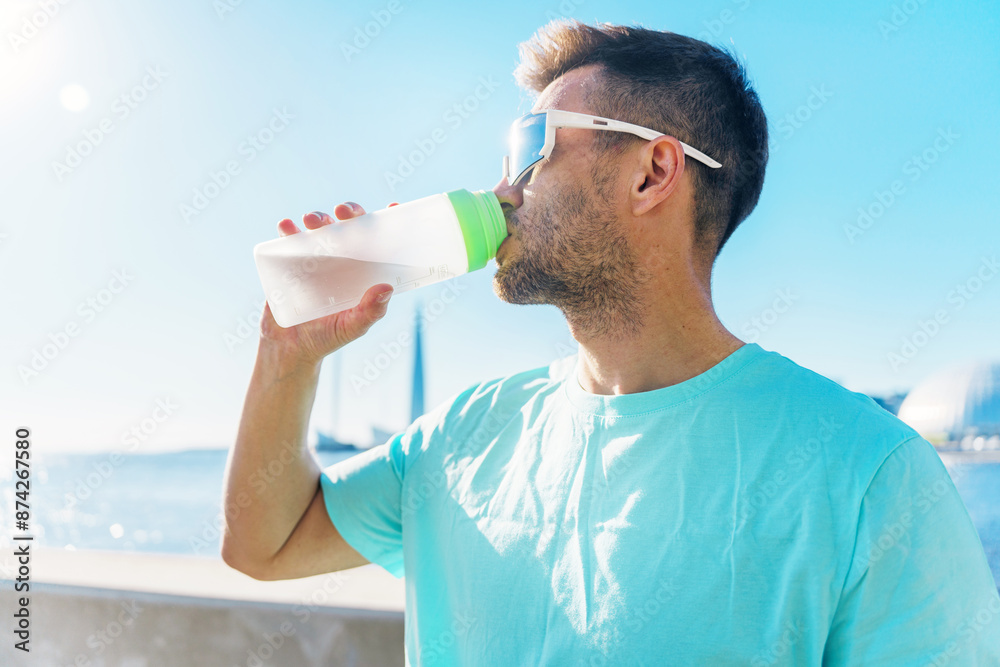 Jogger hydrating with a water bottle, wearing sunglasses, bright sunlight and ocean in the background.