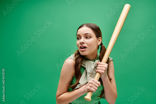 A young woman in her 20s holds a baseball bat confidently in front of a vibrant green background.