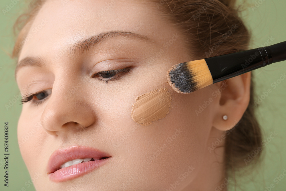 Beautiful young woman with foundation on her face holding makeup brush against green background, closeup Beautiful young woman with foundation on her face holding makeup brush against green background, closeup