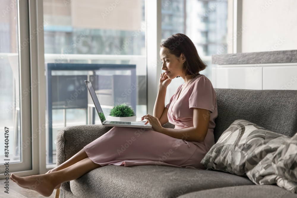 Positive thoughtful young freelancer woman resting on comfortable couch, holding laptop, browsing social media, touching chin, using online app for communication, enjoying movie on Internet Positive thoughtful young freelancer woman resting on comfortable couch, holding laptop, browsing social media, touching chin, using online app for communication, enjoying movie on Internet