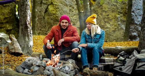 Two young people went hiking in autumn forest. Happy Caucasians spend good time sitting next to bonfire. Man and woman roast marshmallows on fire in the middle of wood. © ihorvsn