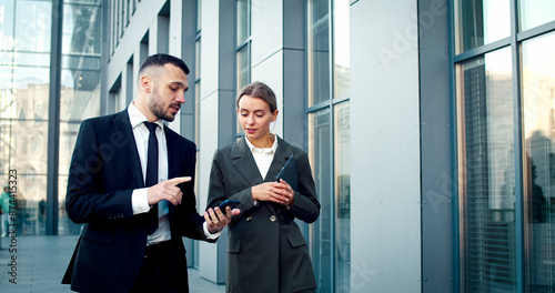 Portrait of Caucasians wearing business suits watch something on phone. Businessman shows female new app on device. Male explains woman how to use mobile application.