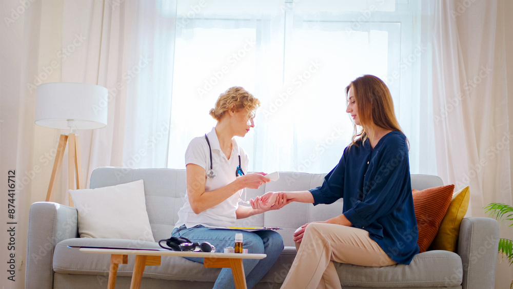 Fototapeta premium Portrait of doctor measures woman's temperature at home. Family doctor examines patient sitting on sofa near table with medications.