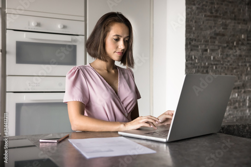 Serious beautiful young freelance entrepreneur girl working at laptop at home, typing on pc on workplace kitchen table, using computer for online job communication