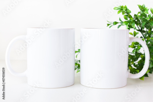 Mockup of two white coffee mugs on table with greenery. Blank 11 oz ceramic cups mock up.