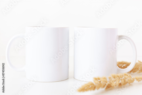 Blank two white coffee mugs on table with dried flowers. 11 oz ceramic cups mockup template in boho style.