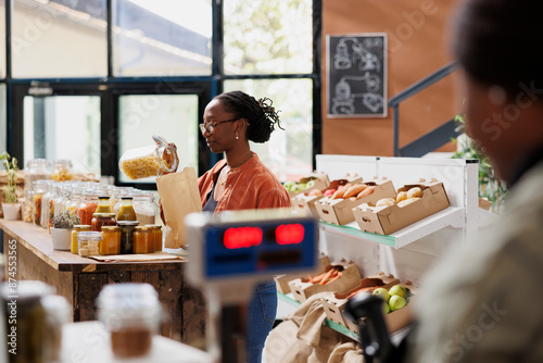 In grocery shop, black woman wearing glasses pours fusilli pasta into bag made of paper. At bio food store, African American female fills a plastic free packaging with freshly made spaghetti. photo