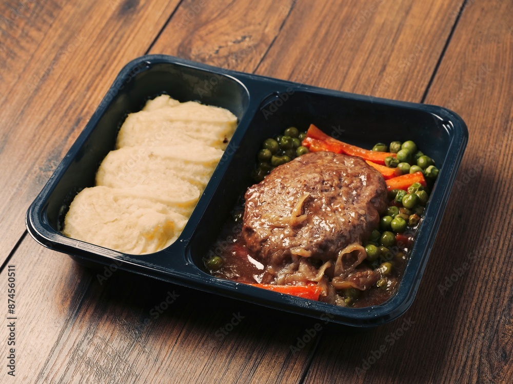 A meal consisting of burger, mashed potatoes and vegetables in plastic tray is served on wooden table and plastic fork and knife. Simple classic dish for take away. Office lunch. A meal consisting of burger, mashed potatoes and vegetables in plastic tray is served on wooden table and plastic fork and knife. Simple classic dish for take away. Office lunch.