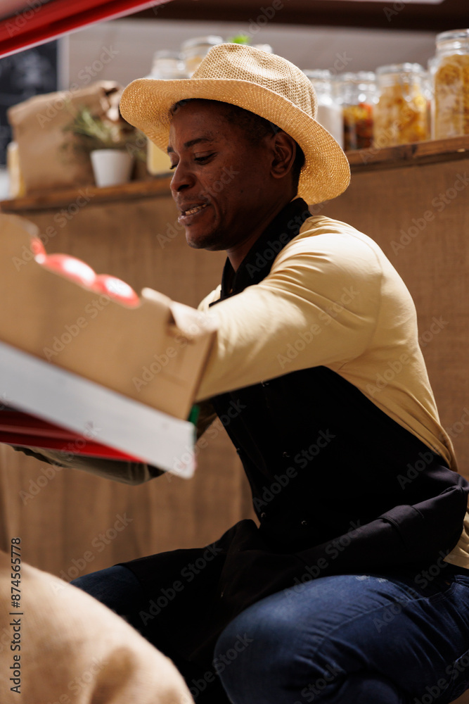 African American business owner arranges eco friendly products in supermarket. Detailed image of a black guy organizing a variety of freshly gathered vegetables on the shelves while wearing an apron. African American business owner arranges eco friendly products in supermarket. Detailed image of a black guy organizing a variety of freshly gathered vegetables on the shelves while wearing an apron.