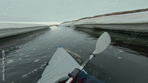 A kayaker paddles through a narrow channel in a frozen river, with snow-covered banks on either side. The sky is overcast, and a flock of birds can be seen flying overhead