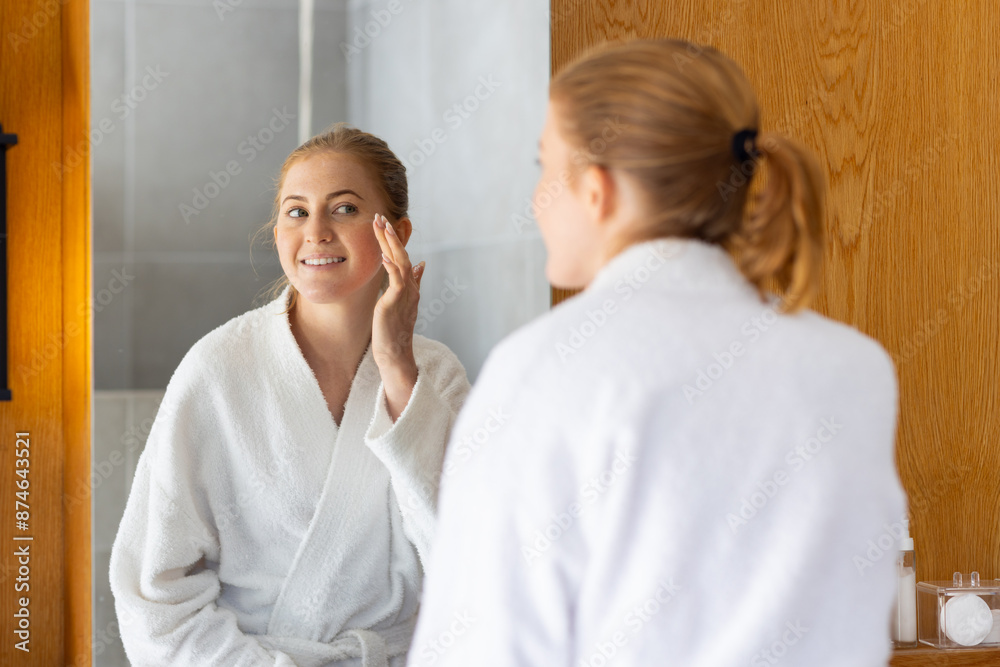 Applying skincare, woman in bathrobe looking in mirror and smiling Applying skincare, woman in bathrobe looking in mirror and smiling