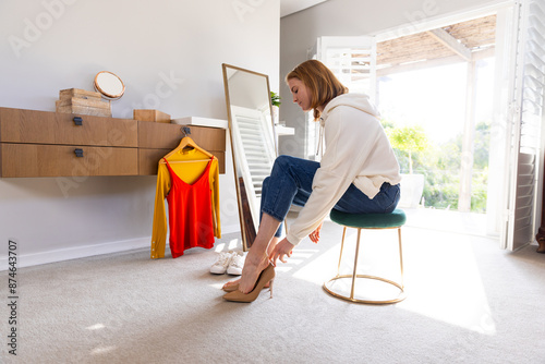 Trying on high heels, woman sitting on stool in bedroom with clothes nearby photo