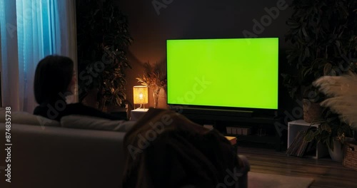 A young lady unwinding after a long day at work by viewing television while seated on a sofa in her comfortable living room, green screen mockup.