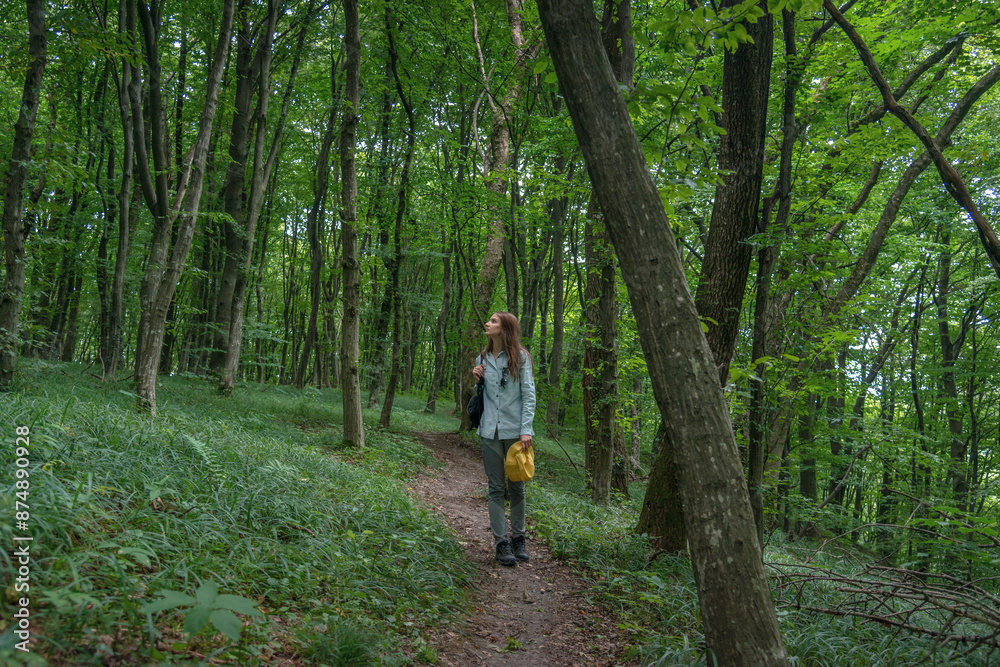 Young Woman Enjoying the Moments of Relaxation in a Forest. Cheerful woman in a yellow hat walking in the deep green forest. Enjoying fresh air and connection with nature. Young Woman Enjoying the Moments of Relaxation in a Forest. Cheerful woman in a yellow hat walking in the deep green forest. Enjoying fresh air and connection with nature.