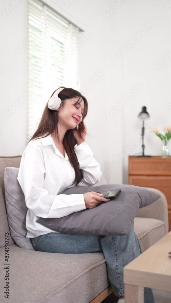 woman is sitting on a couch in a cozy living room, wearing white headphones and smiling as she holds a tablet. She is dressed in a white shirt and appears to be enjoying music or a podcast woman is sitting on a couch in a cozy living room, wearing white headphones and smiling as she holds a tablet. She is dressed in a white shirt and appears to be enjoying music or a podcast