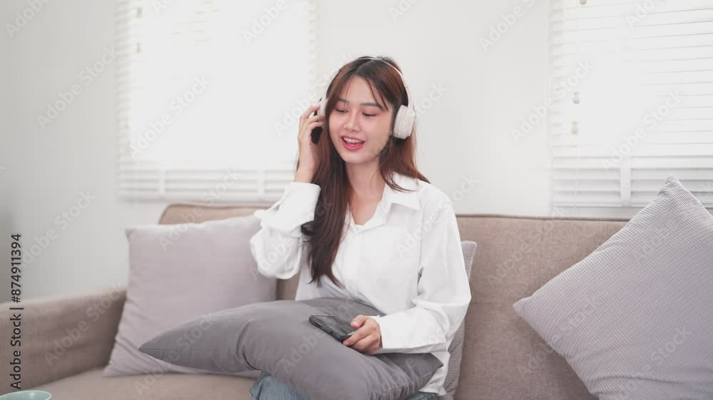 woman is sitting on a couch in a cozy living room, wearing white headphones and smiling as she holds a tablet. She is dressed in a white shirt and appears to be enjoying music or a podcast woman is sitting on a couch in a cozy living room, wearing white headphones and smiling as she holds a tablet. She is dressed in a white shirt and appears to be enjoying music or a podcast