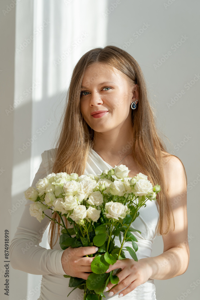 Close portrait of cute bride with long blonde hair wearing white dress and earring holding bouquet of small rose flowers in hands at sunlight looking at camera with relaxed smile at wedding ceremony Close portrait of cute bride with long blonde hair wearing white dress and earring holding bouquet of small rose flowers in hands at sunlight looking at camera with relaxed smile at wedding ceremony