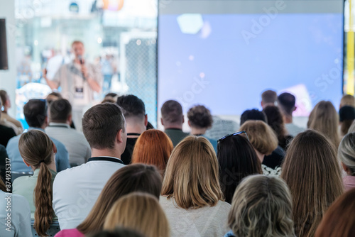 Large group of people attending a business conference, focusing on a speaker presenting on stage. Professional event with business attire and engaged audience.