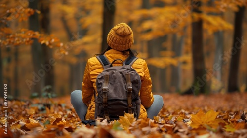 Person wearing a yellow jacket and beanie sitting in a forest surrounded by golden autumn leaves, enjoying the serene fall scenery.