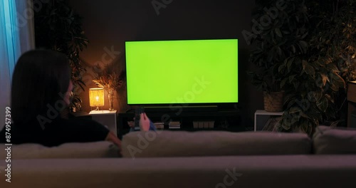 A young woman sitting on couch in the cozy living room watching a television whileresting relaxing after hard long day at work at home green screen mockup.
