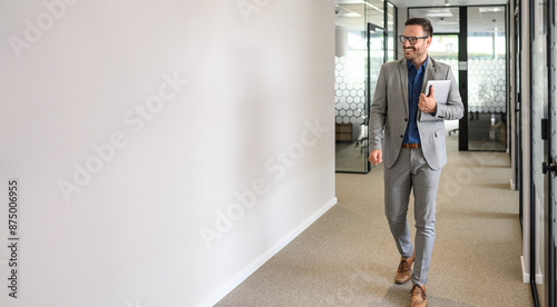 Full length of happy male leader with laptop looking away and walking by white wall in office corridor