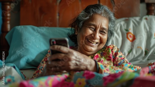 a senior smiling indian woman lying in bed, looking at her cell phone