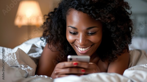 a young smiling african, american woman lying in bed, looking at her cell phone