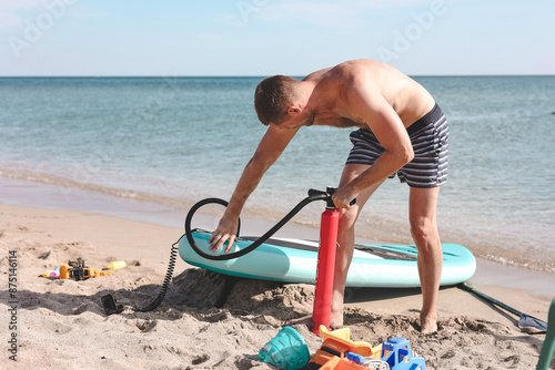 Man inflating stand up paddle board with manual pump at sea background. Middle-aged man preparing blue SUPboard for surfing.