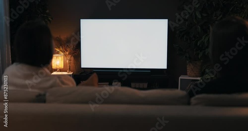 Two friends are talking last gossips weekends concept. Happy women sitting in the living room watching tv news movie white screen mockup television in cozy living room over the shoulder shot.