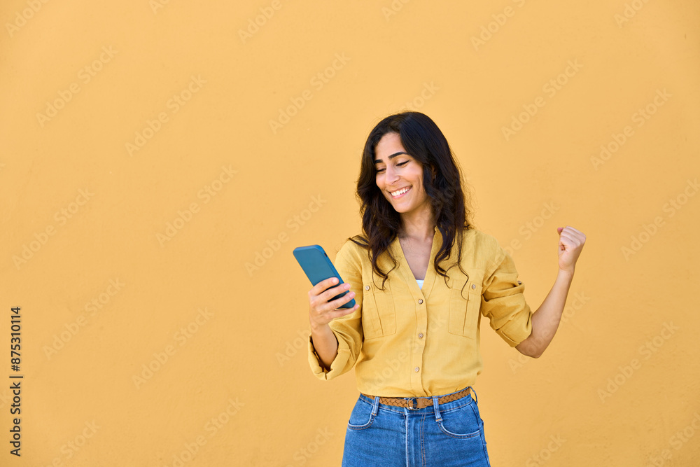 Excited happy young Arabian or Indian woman shopper winner holding cell phone isolated on yellow wall. Middle eastern female student feeling joy using mobile app, winning gift, bonus, reading news Excited happy young Arabian or Indian woman shopper winner holding cell phone isolated on yellow wall. Middle eastern female student feeling joy using mobile app, winning gift, bonus, reading news