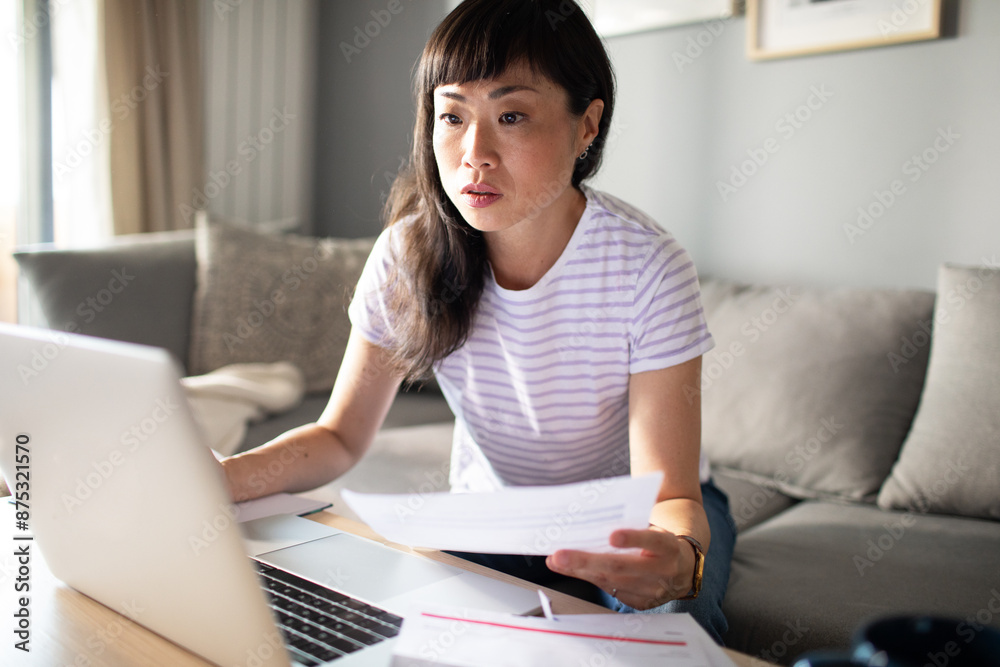 Concerned Asian woman holding bills and using laptop at home Concerned Asian woman holding bills and using laptop at home