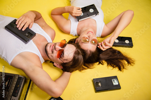 A woman and a man in sunglasses are lying on a yellow background. An old tape recorder and video cassettes are nearby. Retro style, music.