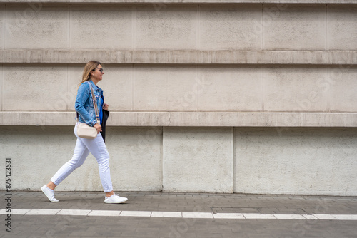 Young slim woman in white pants and denim katana walking with black bag along the sidewalk in the city against the background of gray building. Everyday life in city. Side view. © Jacek Chabraszewski