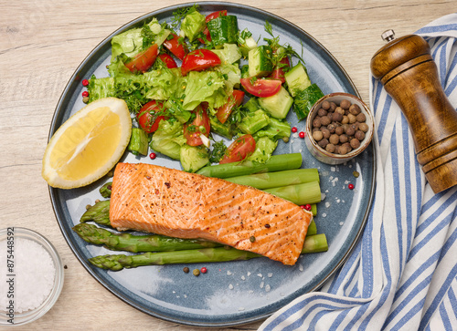 Healthy lunch with grilled salmon with asparagus and fresh tomato and cucumber salad, a row of lemon slice.