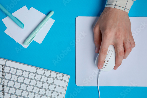 Man working with mouse, computer keyboard and stationery at light blue table, top view